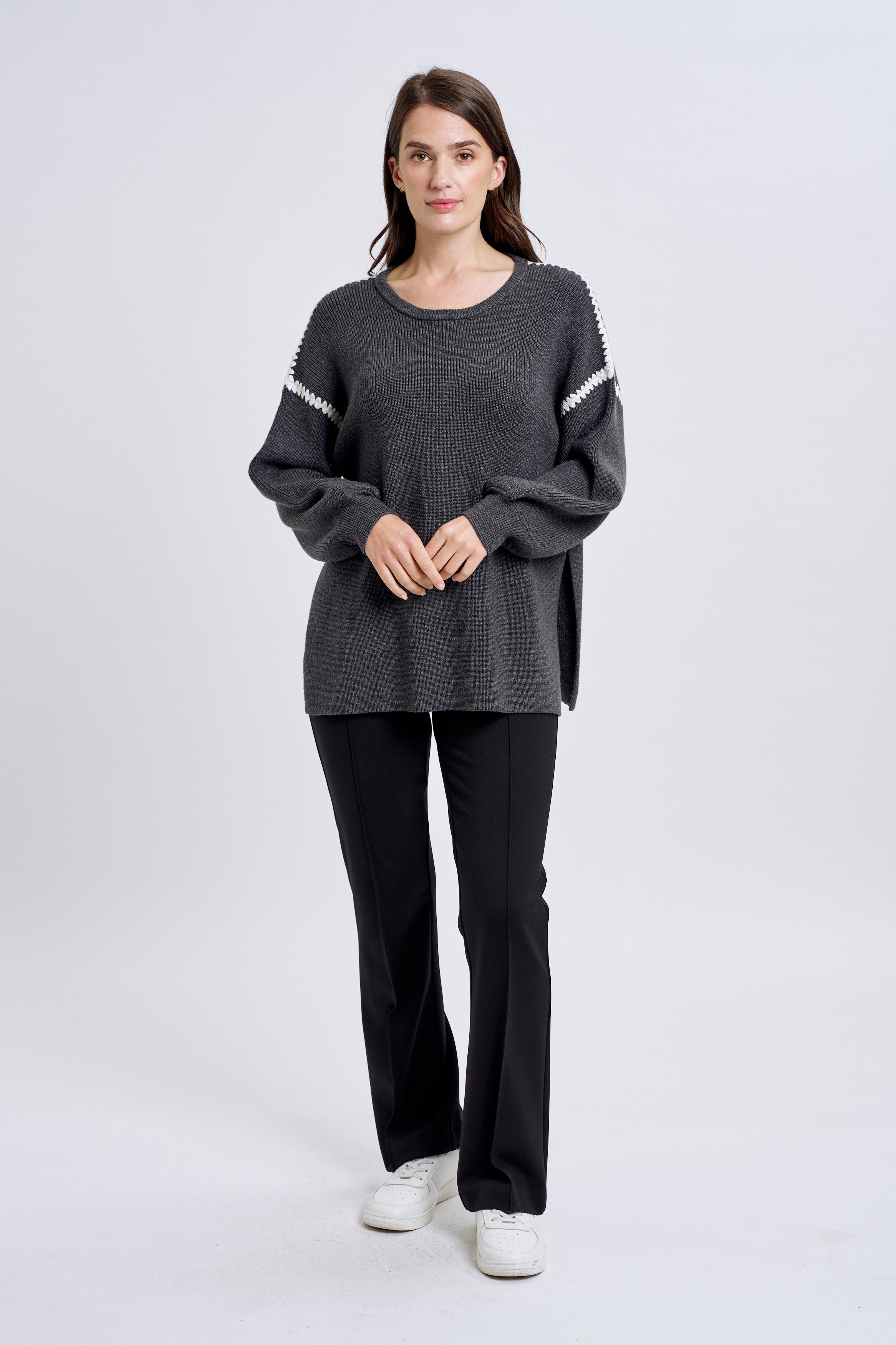 A female model wearing a gray, long-sleeved, knitted sweater with blanket stitch detailing and side slits. She is also wearing black pants and white shoes.