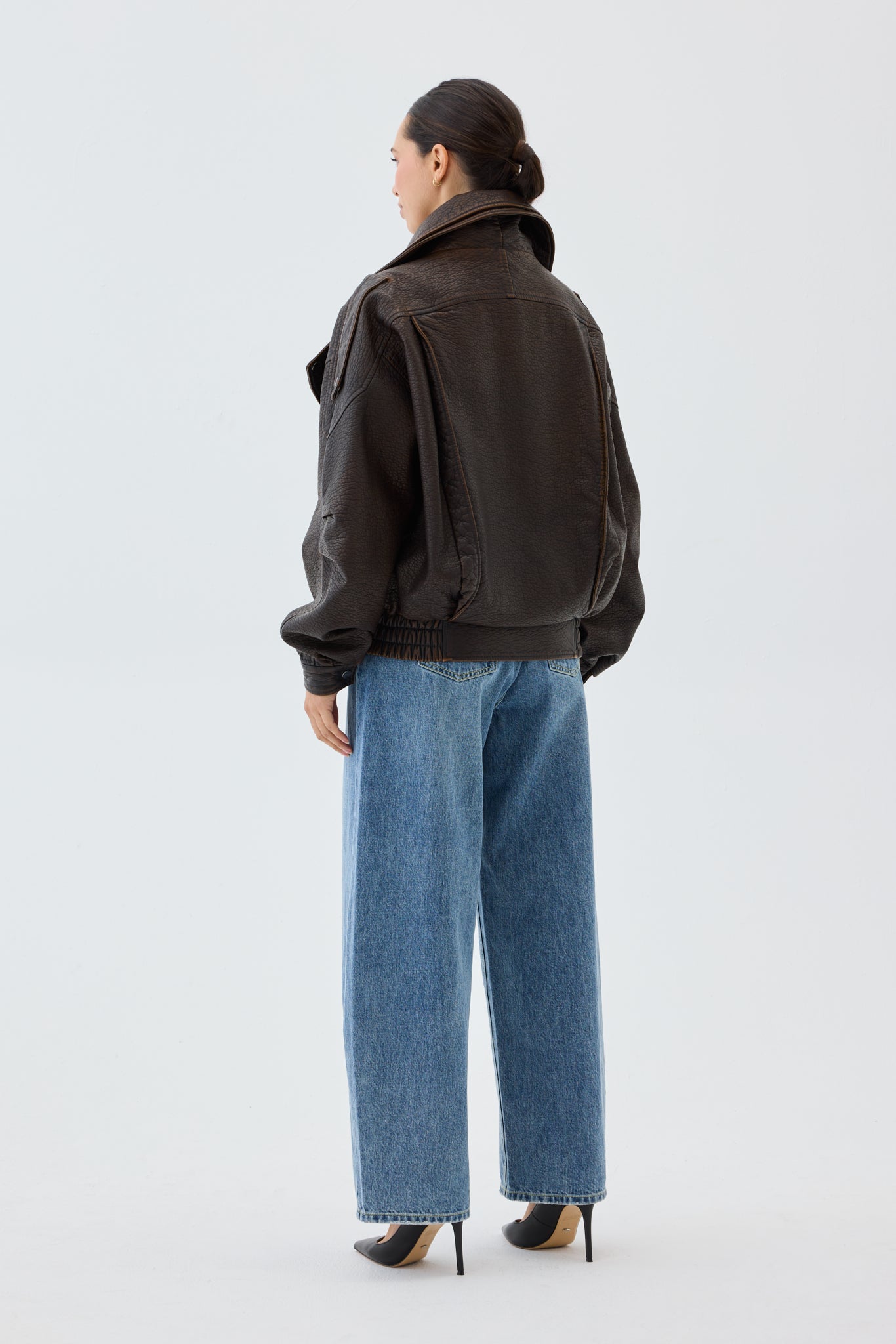 Woman wearing an oversized, brown faux leather bomber jacket and blue jeans with a white background.