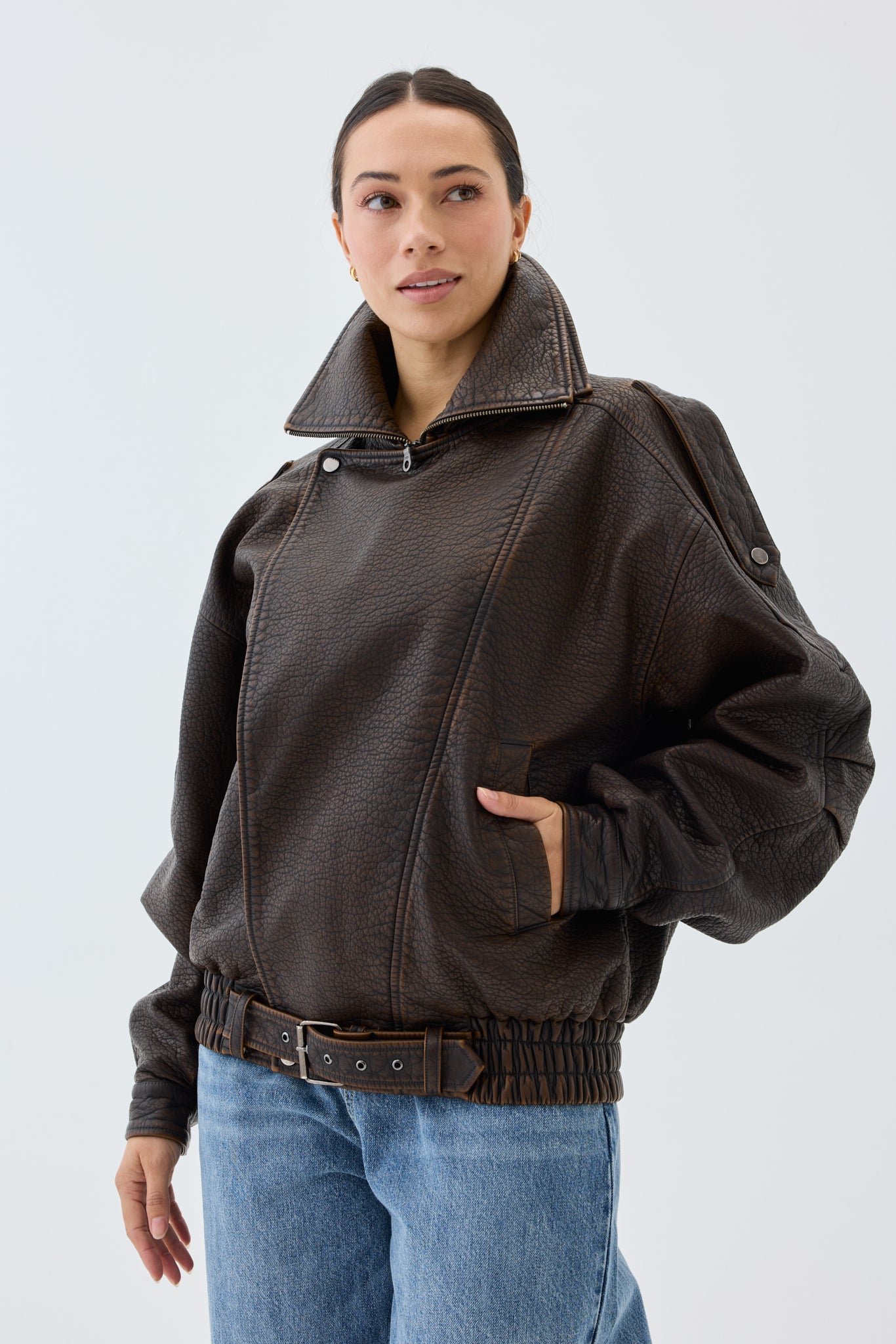 Woman wearing an oversized, brown faux leather bomber jacket and blue jeans with a white background.