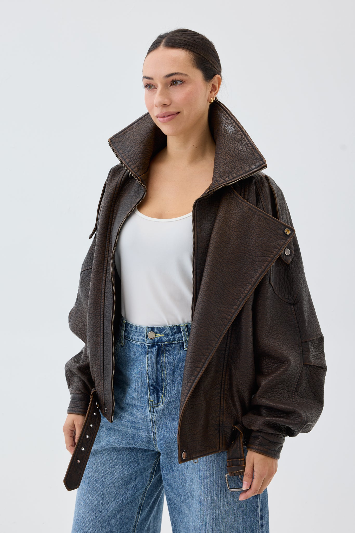 Woman wearing an oversized, brown faux leather bomber jacket with white top and blue jeans in front of a white background.