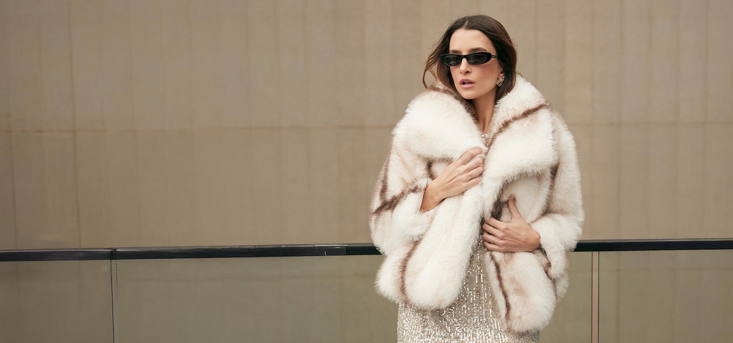 Woman wearing a white fur coat and sunglasses against a beige wall.