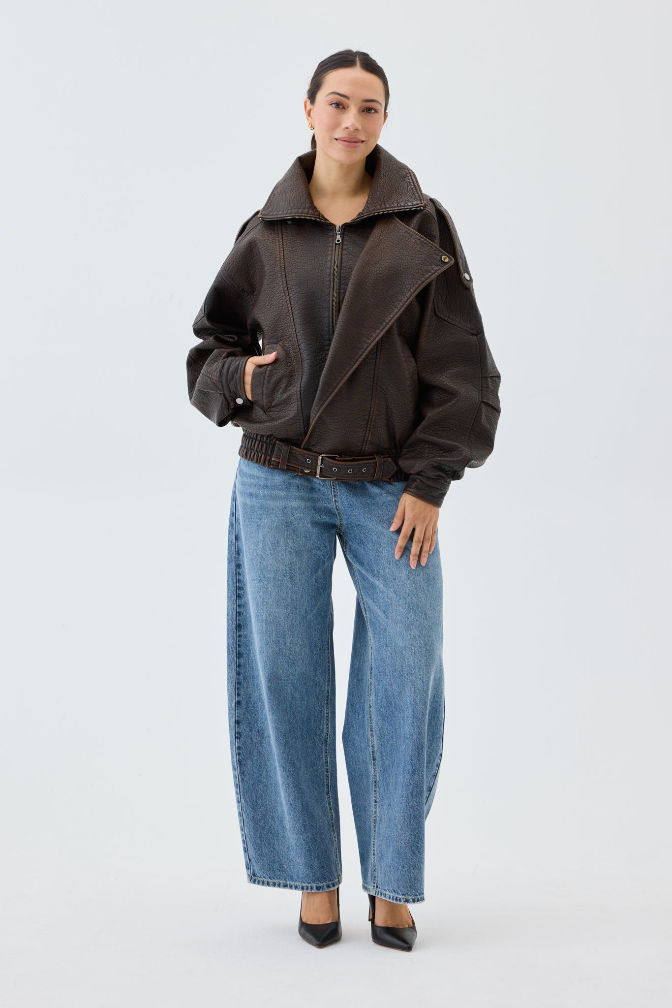 Woman wearing an oversized, brown faux leather bomber jacket and blue jeans with a white background.